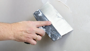 A hand holding a broad taping knife to apply joint compound onto a smooth wall.