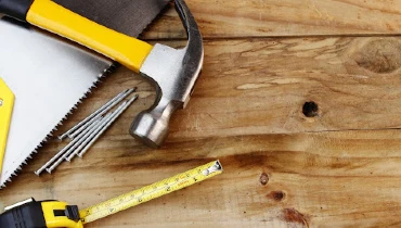 A hammer, saw, tape measurer, and nails laying on top of wooden planks before they’re used for floor repair.