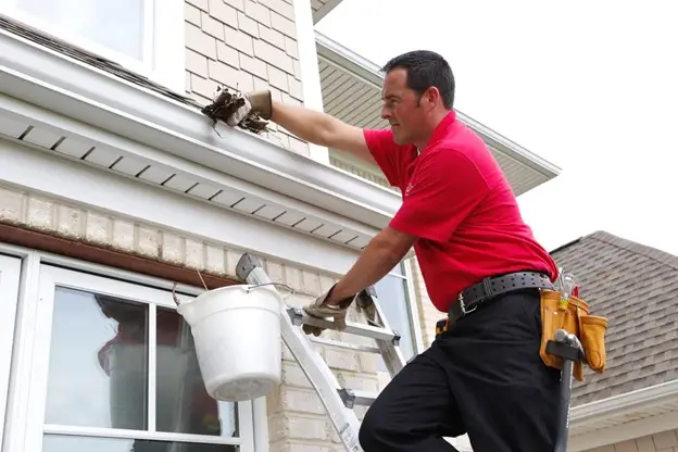 A handyman from Mr. Handyman standing on a ladder and reaching into a home’s gutters as he provides service for gutter cleaning in Collinsville, IL.