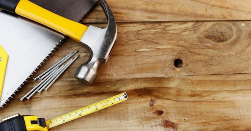 A hammer, saw, tape measurer, and nails laying on top of wooden planks before they&rsquo;re used for floor repair.