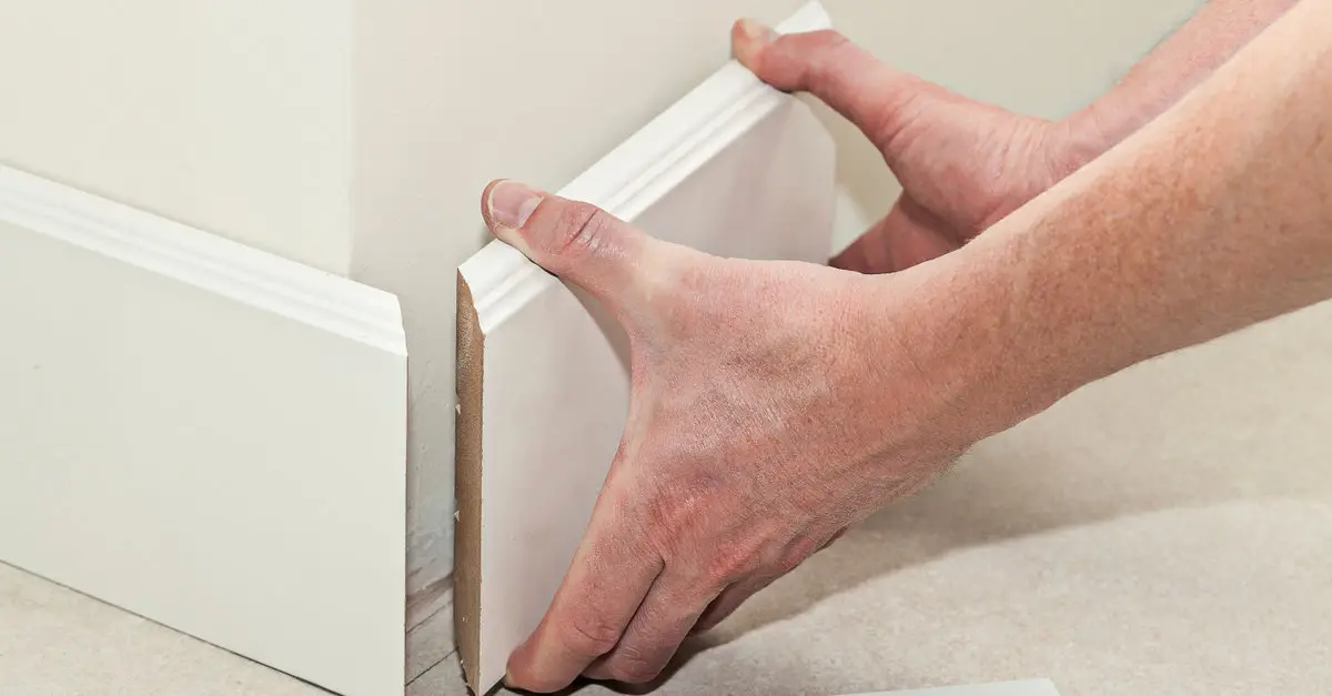 A close up of a handyman&rsquo;s hands as he installs a new section of baseboard trim along the base of a wall.
