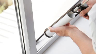 A person applying sealant to the gap between a window frame and the glass using a caulking gun.